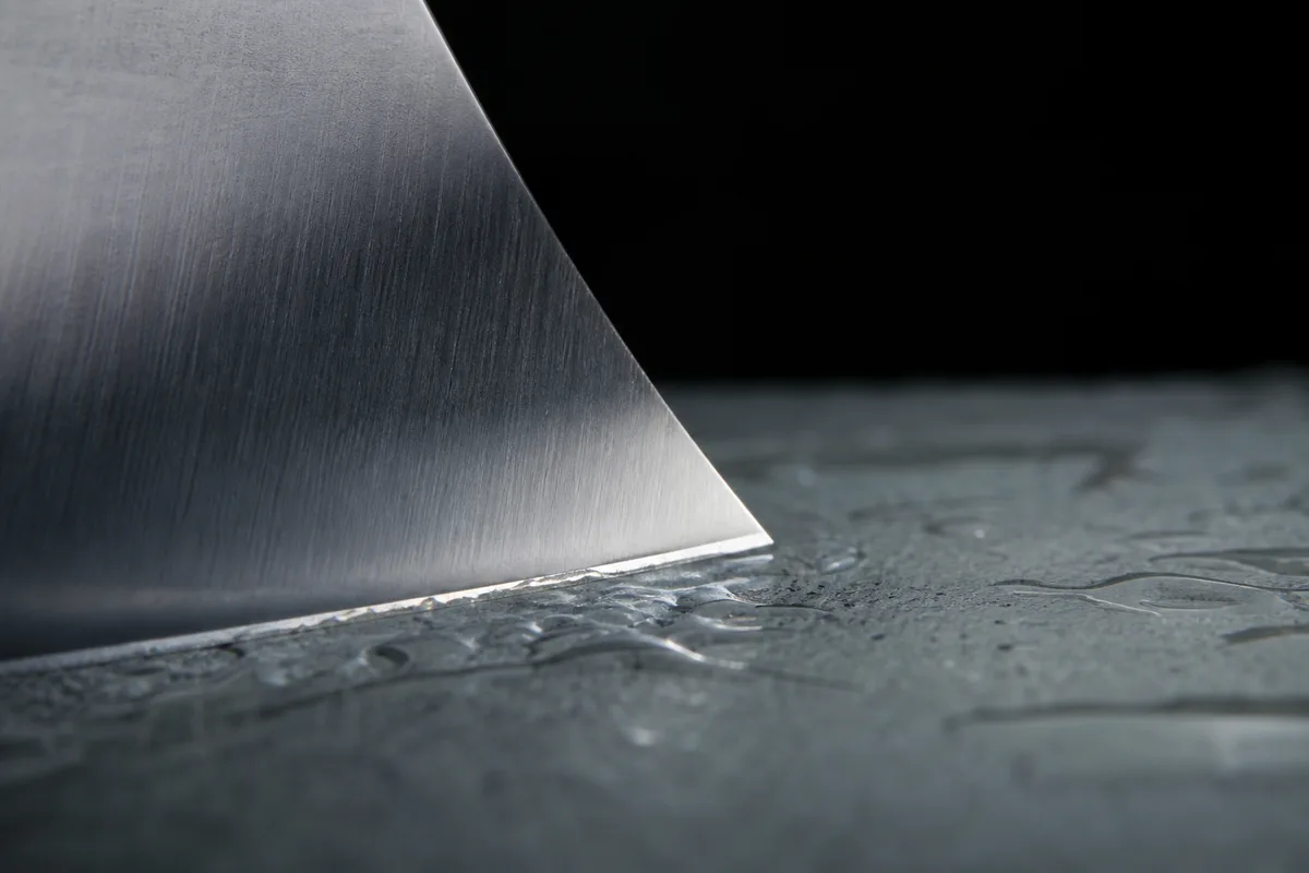 Gros plan sur le fil d une lame de couteau de chef posée sur une pierre à eau, boue abrasive grise, lumière rasante